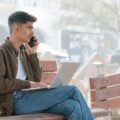 A young man multitasking with a laptop and smartphone on a park bench, embracing remote work outdoors.